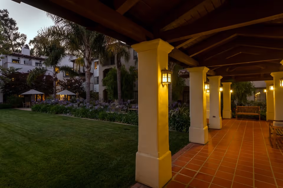 Covered outdoor walkway with tiled floor and columns lit by wall-mounted lanterns, adjacent to a well-maintained lawn and garden area with palm trees and umbrellas near a multi-story building at dusk.