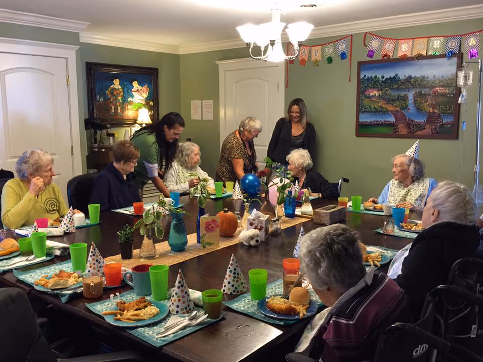A group of elderly people and two caregivers gathered around a large dining table in a decorated room celebrating a birthday. The table is set with plates of food, colorful cups, party hats, and small plants. A birthday banner and a painting are visible on the green walls.