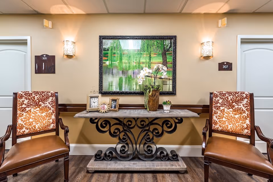 A hallway area in a senior living facility with two wooden chairs featuring orange and white patterned upholstery. Between the chairs is a decorative table with a wrought iron base and a wooden top, holding framed photos and a vase with white flowers. Above the table hangs a large framed painting of a serene pond with trees. Two wall sconces provide lighting, and there are two white doors on either side labeled 305 and 307.