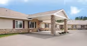 Front exterior of a single-story senior living facility with a covered entrance/porte-cochère, stone columns, and driveway.