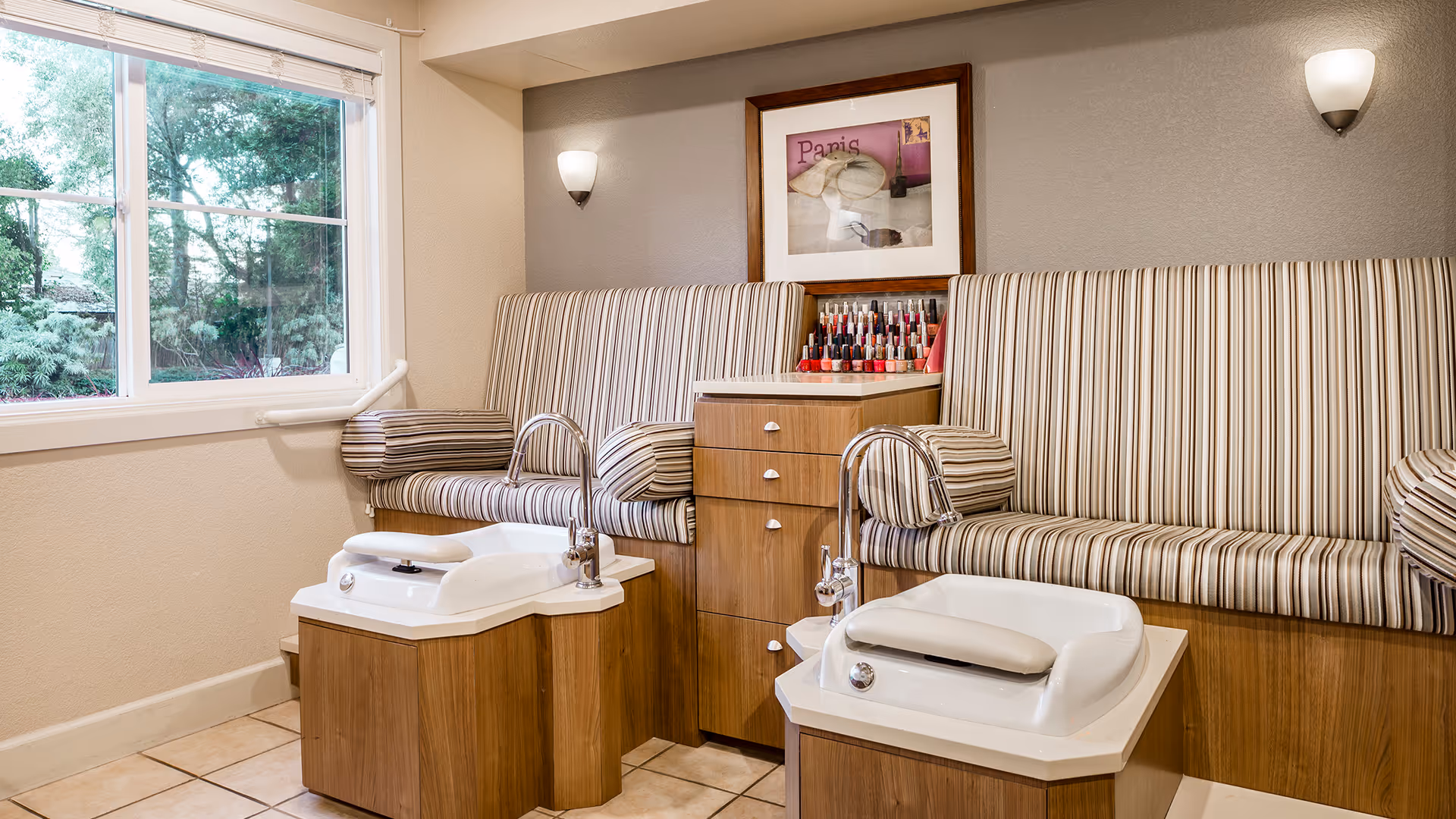 A cozy nail salon area with two striped cushioned benches and two white pedicure foot baths with faucets. There is a wooden cabinet between the benches holding various nail polish bottles. A large window shows greenery outside, and two wall-mounted lights illuminate the room.