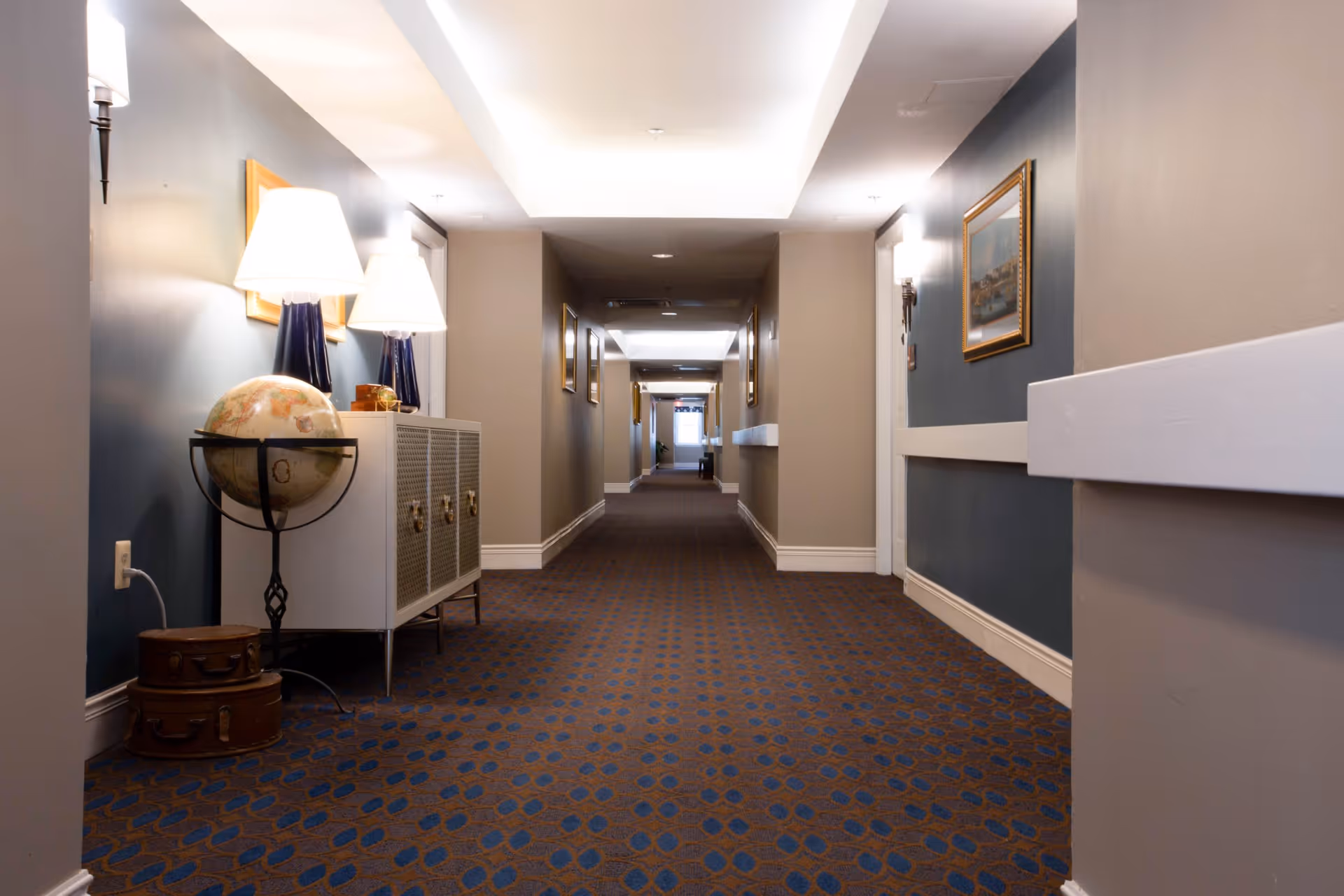 A long, carpeted hallway in a senior living facility with blue and brown patterned carpet, beige walls, and white trim. The hallway is decorated with framed paintings on the walls and has a small cabinet with two lamps and a globe on the left side. The ceiling has recessed lighting and a white tray ceiling design.