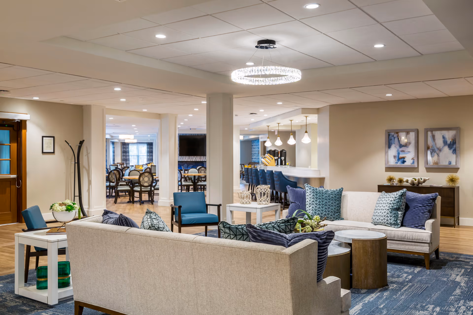 A spacious and well-lit senior living facility common area with beige sofas adorned with blue and patterned throw pillows, a blue armchair, and round wooden coffee tables. In the background, there is a dining area with multiple tables and chairs, and a bar counter with blue bar stools and hanging pendant lights. The walls are decorated with abstract artwork and the floor has a combination of wood and blue patterned carpet.