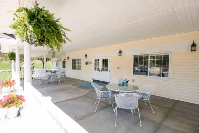 Covered outdoor patio with round metal tables and chairs, hanging ferns, and potted flowers along a light-colored building.