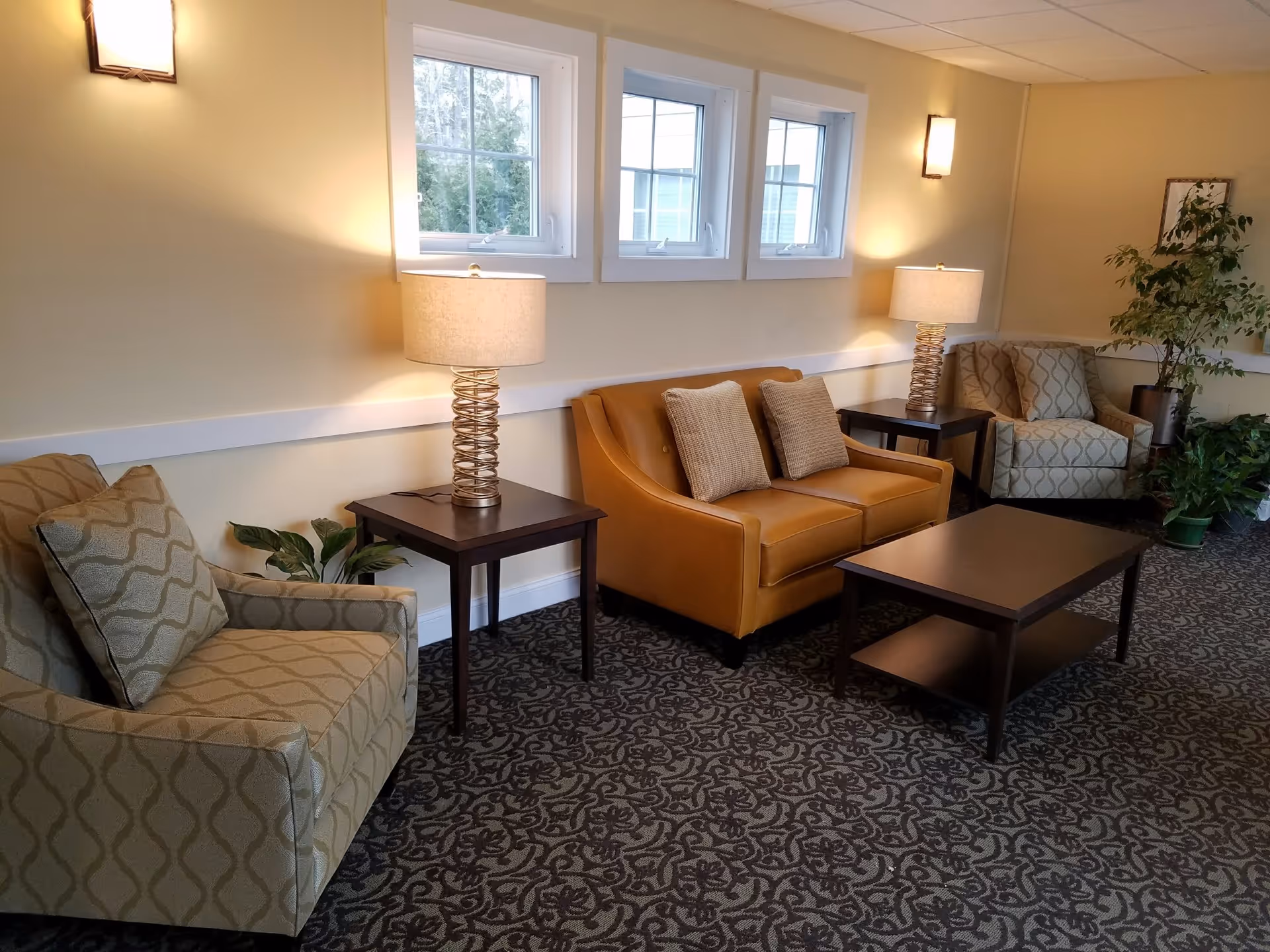 A cozy living room area with two patterned armchairs, a tan leather loveseat with two cushions, two side tables each with a lamp, a rectangular coffee table, and several potted plants. The walls are light-colored with three windows letting in natural light.