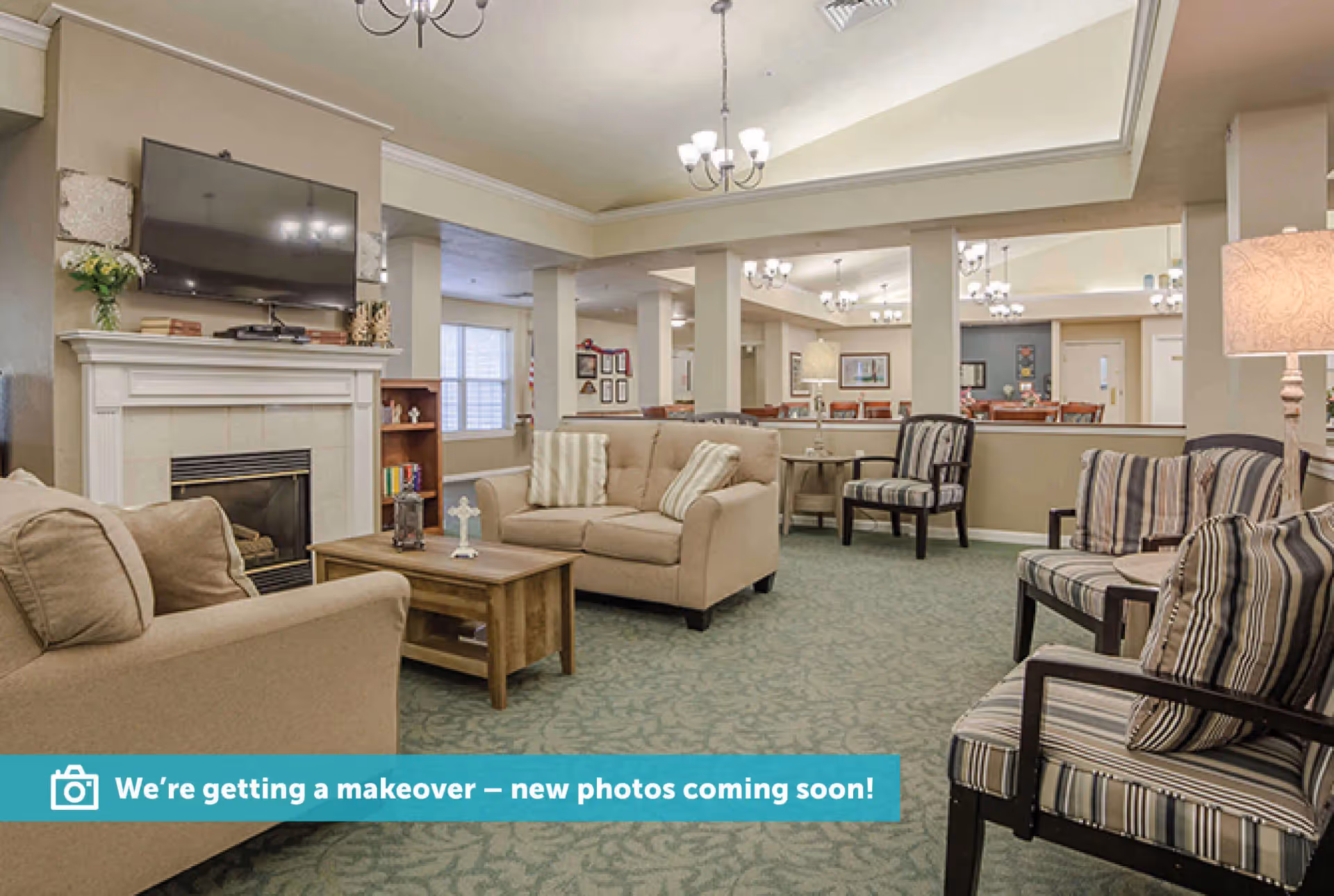 A spacious living room area with beige sofas, striped armchairs, a wooden coffee table, a fireplace with a mounted flat-screen TV above it, and several chandeliers hanging from the ceiling. The room has carpeted flooring and is decorated with lamps, framed pictures, and a bookshelf.