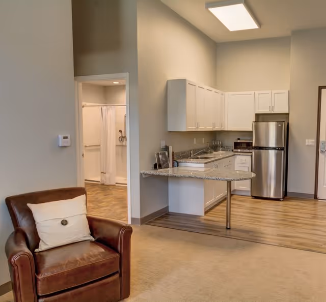 Interior view of a senior living facility showing a small kitchen area with white cabinets, a granite countertop, a stainless steel refrigerator, and a toaster oven. To the left, there is a brown leather armchair with a white pillow. In the background, an open doorway reveals a bathroom with a shower and a white shower curtain.