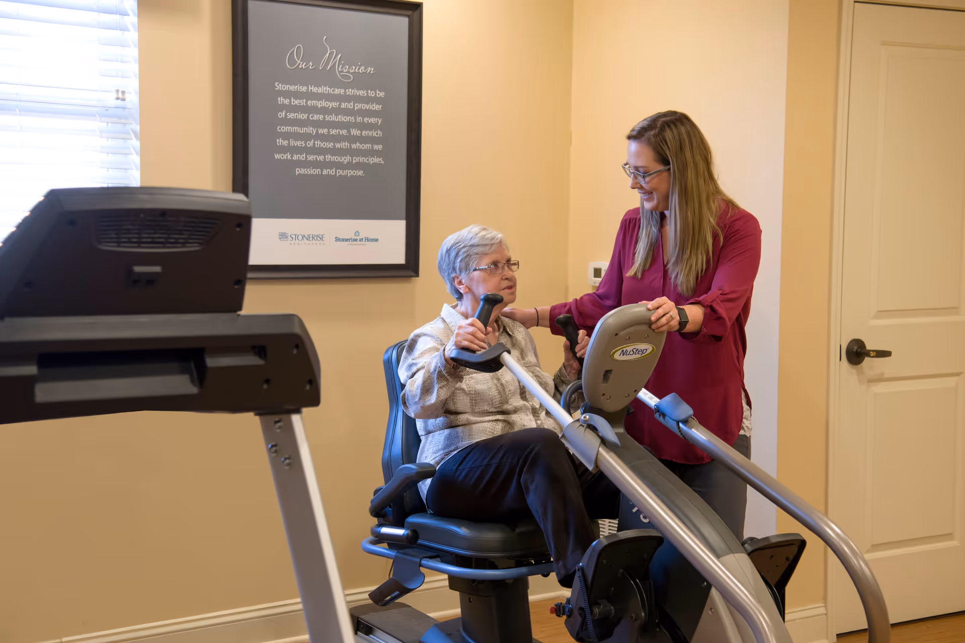 An elderly woman using a NuStep exercise machine with the assistance of a smiling caregiver in a room with beige walls and a framed mission statement on the wall.