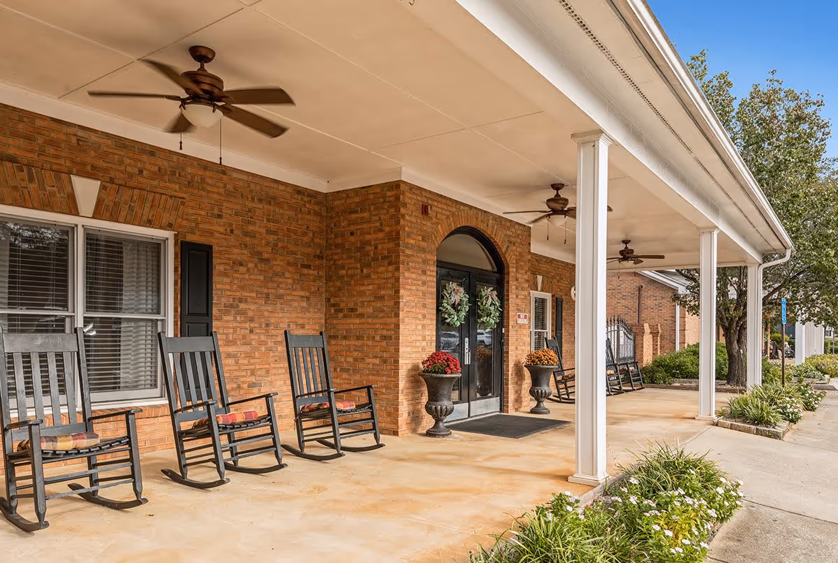 Covered porch area of a brick building with several black rocking chairs with cushions, ceiling fans, potted plants, and double glass doors decorated with wreaths. There are white columns supporting the porch roof and landscaping with green plants and flowers along the walkway.