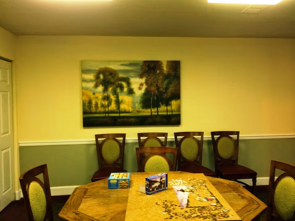 A communal room with a wooden table holding a partially completed jigsaw puzzle and puzzle boxes, surrounded by chairs and a landscape painting on the wall.