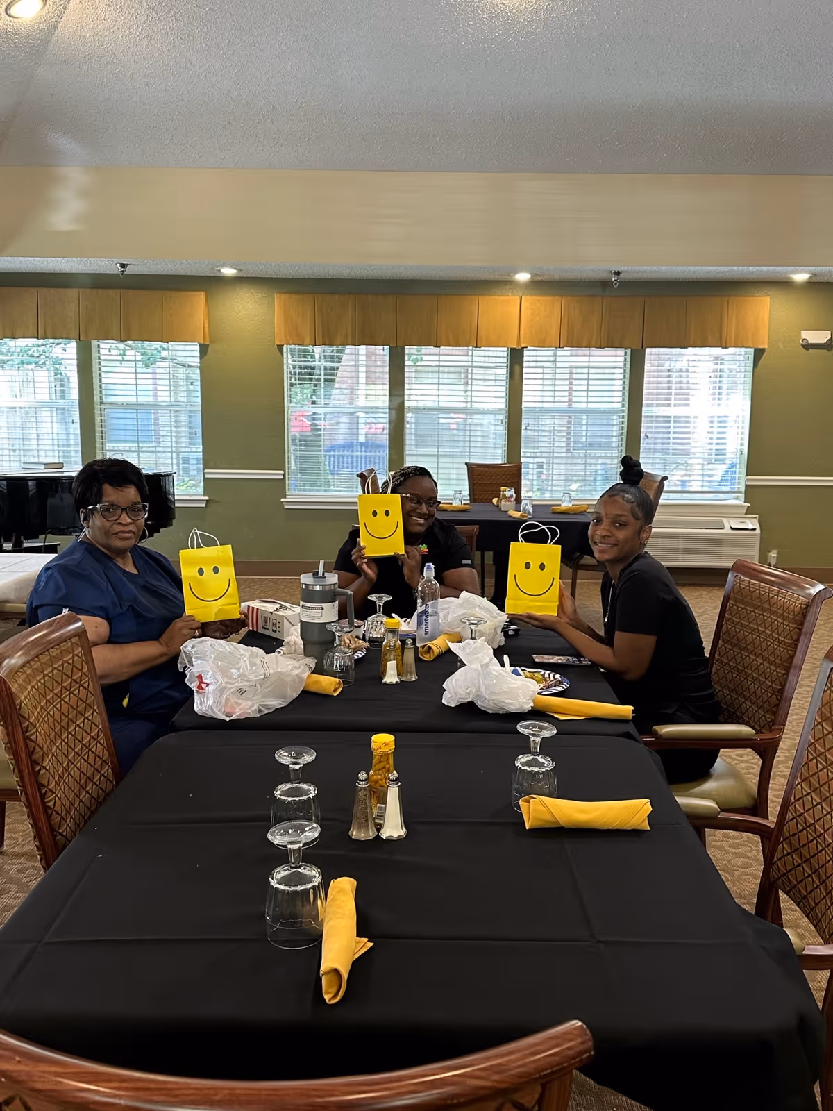 Three women sitting at a dining table in a senior living facility, each holding a yellow gift bag with a smiley face. The table is set with black tablecloth, yellow napkins, upside-down glasses, and condiments. Large windows with blinds and valances are in the background.