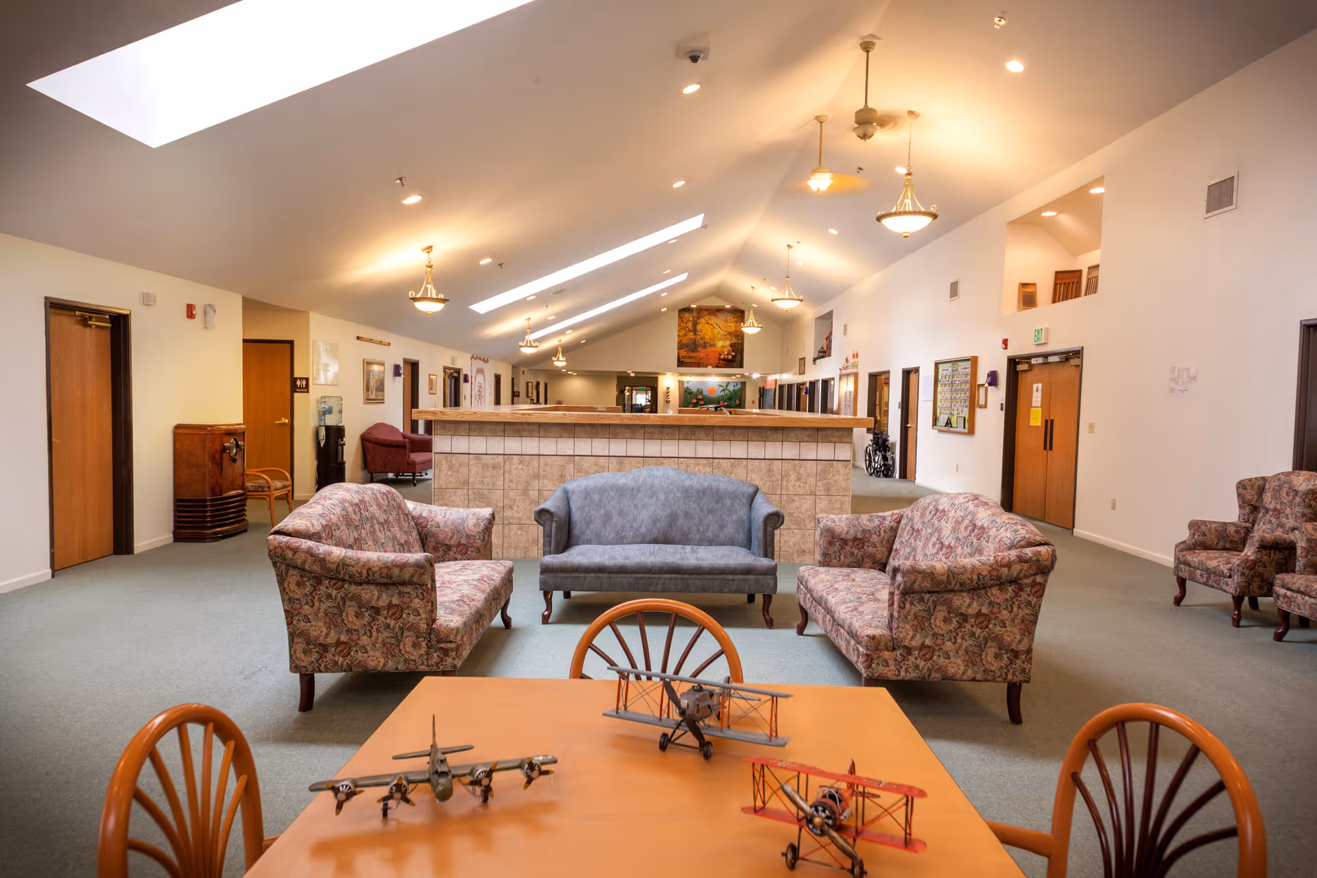 A spacious and well-lit common area in Walnut Ridge Memory Care featuring a high vaulted ceiling with skylights and hanging light fixtures. The room has a seating arrangement with two floral-patterned sofas and one blue sofa facing a tiled half-wall. In the foreground, there is a wooden table with three model airplanes and wooden chairs. The walls are white with several doors and framed pictures, and the floor is carpeted in green.