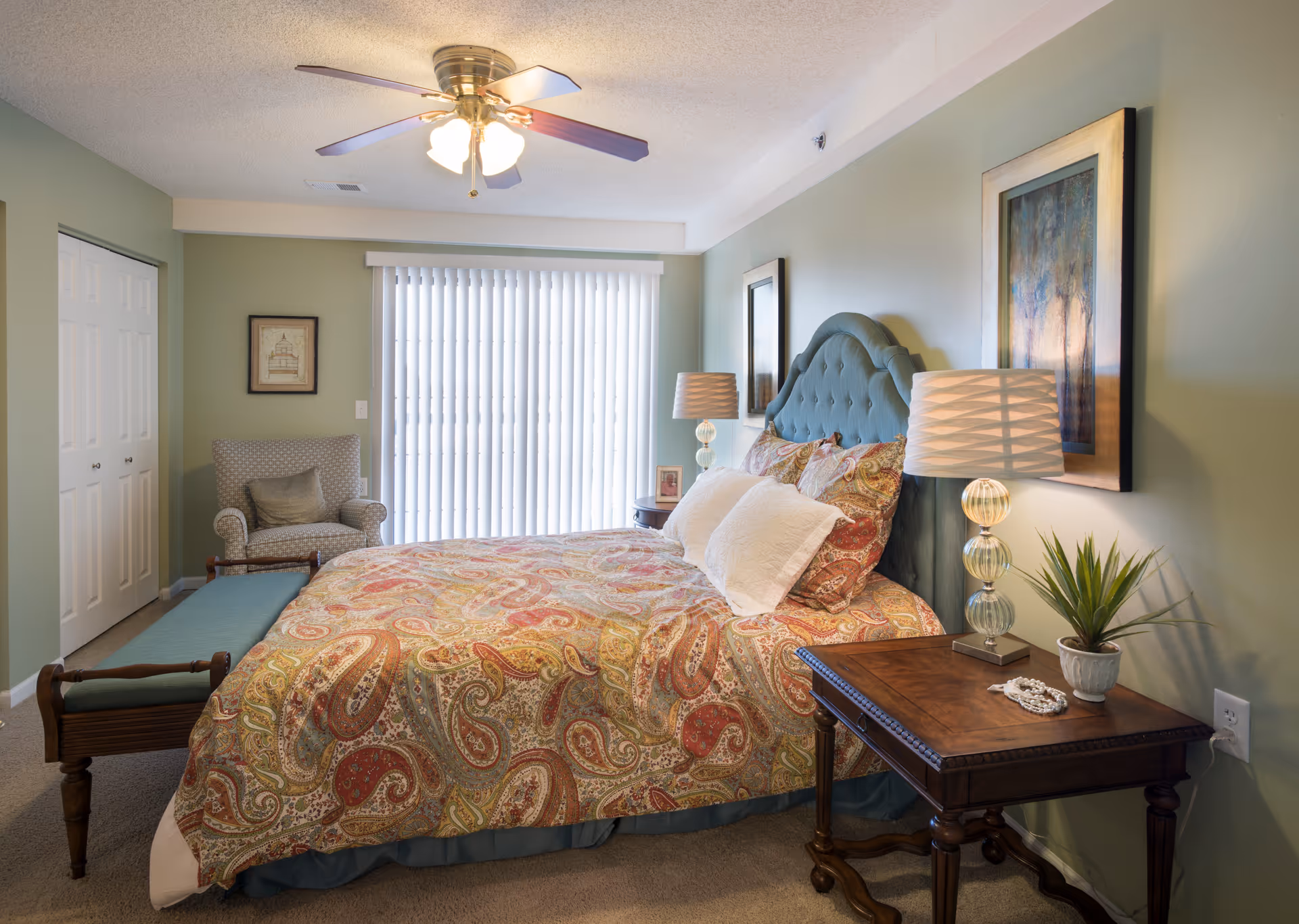 Bright bedroom featuring a patterned quilt on a bed with a teal tufted headboard, bedside tables with lamps, an armchair, bench, ceiling fan, and vertical blinds.