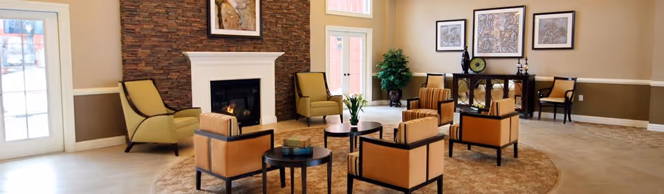 A spacious living room area with a stone fireplace in the center, surrounded by several cushioned chairs and small round tables. The room has large windows and glass doors letting in natural light, beige walls with white trim, and decorative framed artwork above a console table against the far wall. There is a potted plant near the windows adding greenery to the space.
