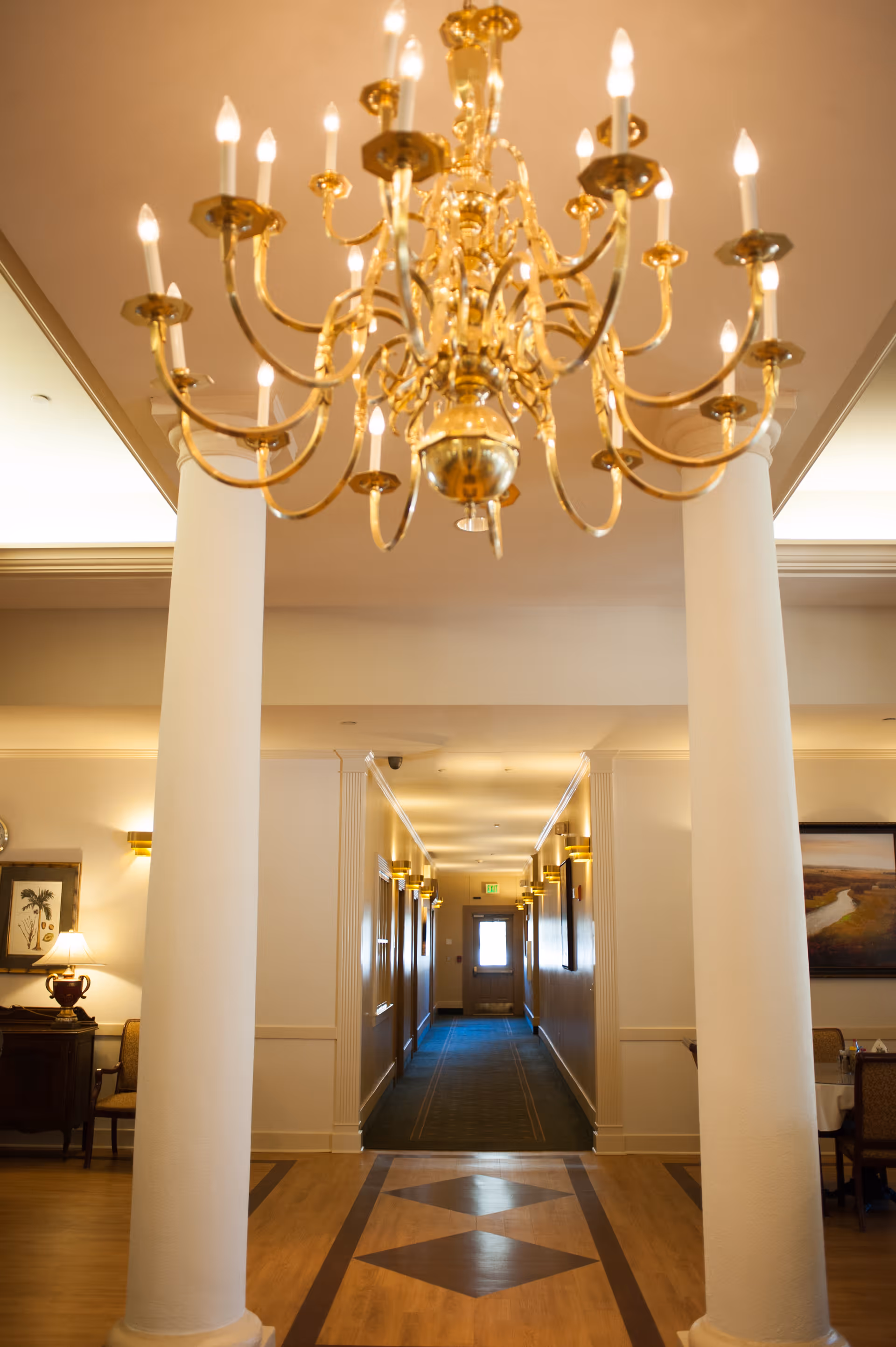 Interior view of a hallway in a senior living facility with a large ornate gold chandelier hanging from the ceiling. The hallway is flanked by white columns and leads to a door at the far end. The floor has a patterned wood design, and there are wall sconces providing warm lighting along the corridor. On the sides, there are chairs, tables, and framed artwork on the walls.