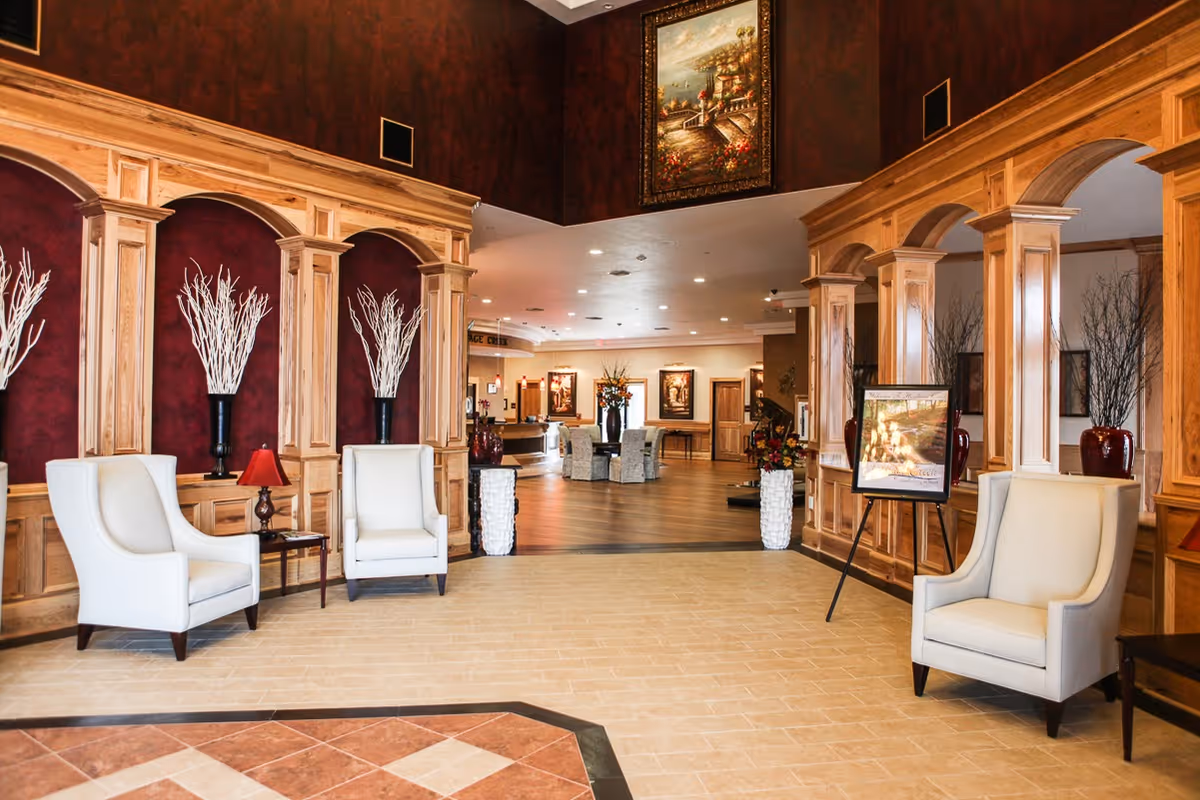 A spacious and elegant interior lobby area with beige tiled flooring and wooden archways. There are three white armchairs arranged near the archways, with decorative vases containing white branches placed in niches along the walls. In the background, a seating area with a round table and chairs is visible, along with framed artwork on the walls and warm lighting.