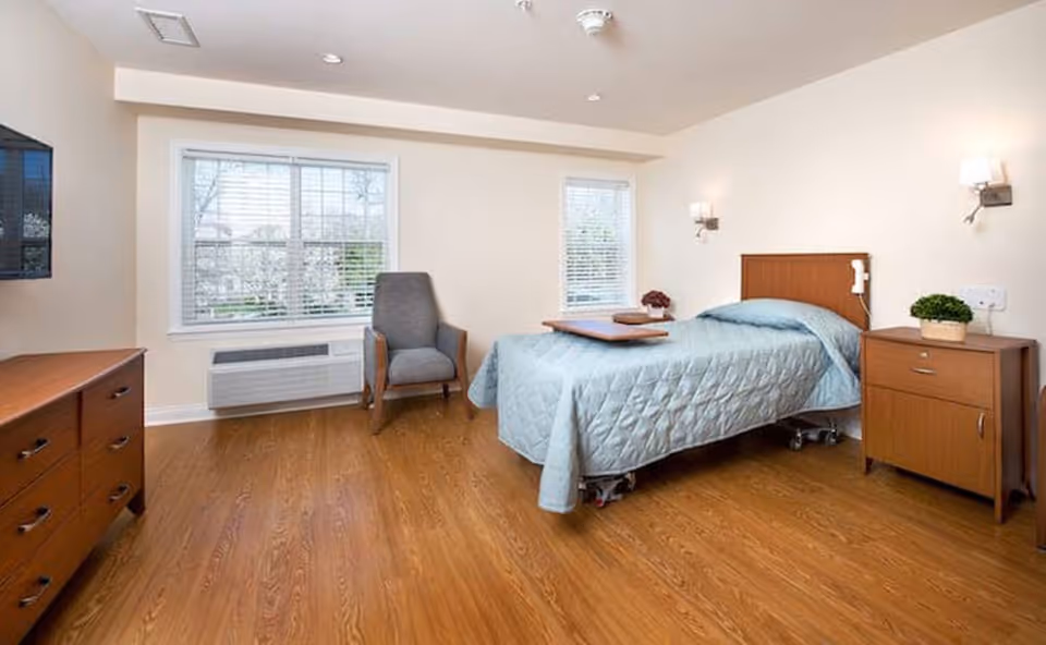 A clean and bright bedroom in a senior living facility with a single bed covered in a light blue quilt, a wooden nightstand with a small plant, a wooden dresser, a gray armchair, and two windows with white blinds letting in natural light. The floor is wood, and the walls are painted white.