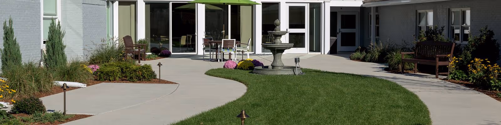 Outdoor courtyard area with a curved concrete walkway, green grass, a central water fountain, patio tables with chairs under a green umbrella, and benches along the building walls surrounded by plants and flowers.