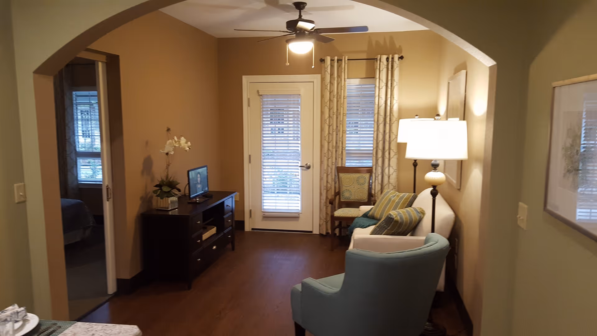 Interior living room featuring an arched entry, a sofa and blue armchair, a TV on a media console, floor lamp and a glass door with blinds.