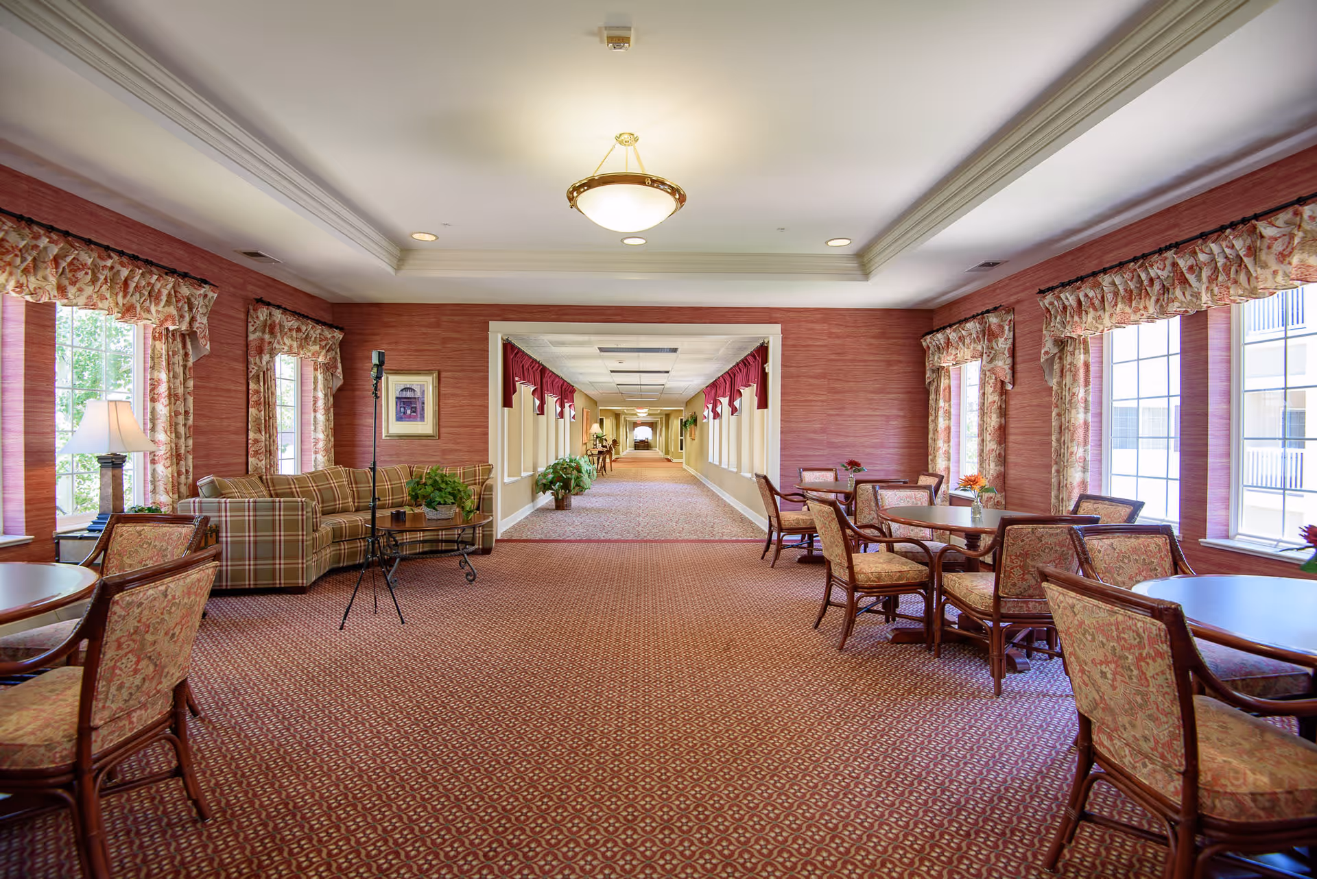 Bright communal lounge and dining area with tables and chairs, a sofa, patterned carpet, and a long central hallway.