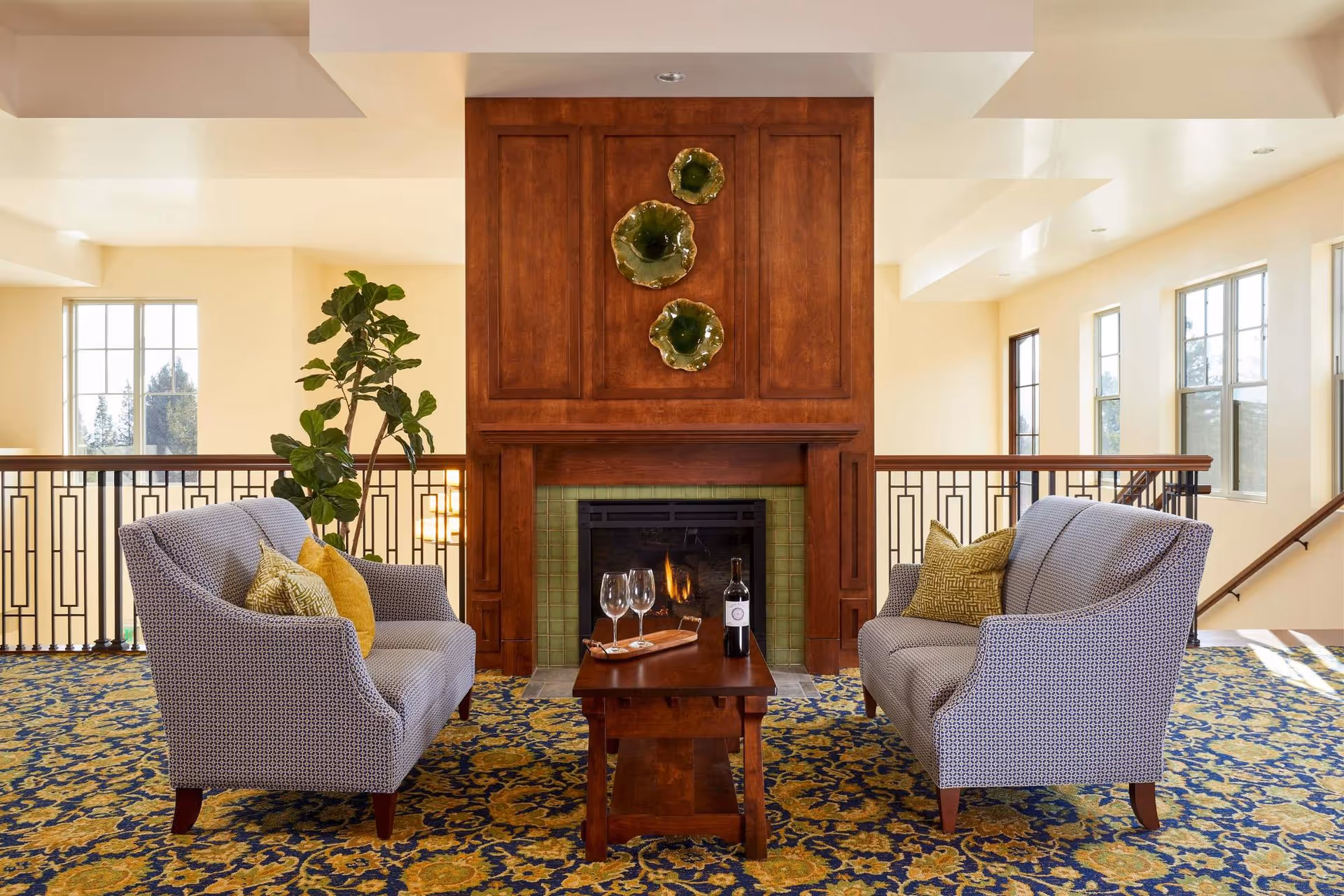 A cozy sitting area with two patterned sofas facing each other across a wooden coffee table holding a bottle of wine and three wine glasses. Behind the table is a fireplace with a wooden mantel and green tile surround, decorated with three green ceramic wall hangings. The space has large windows letting in natural light, a patterned carpet, and a potted plant beside one sofa.