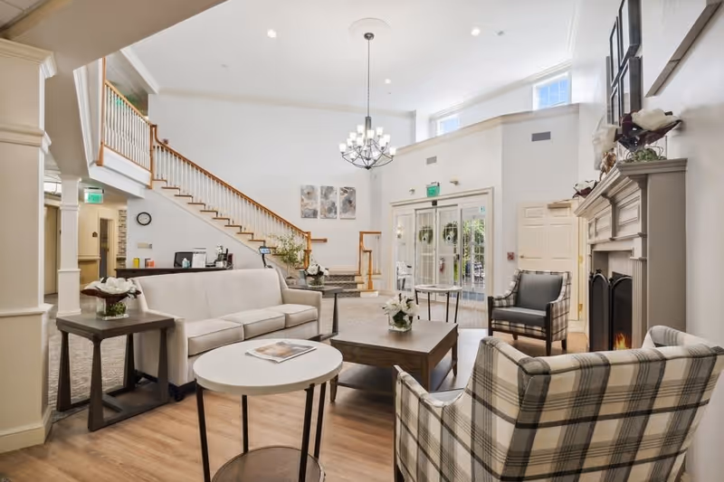 A bright and spacious senior living facility common area with a beige sofa, plaid armchairs, a wooden coffee table, and a fireplace. There is a staircase with wooden railings, a chandelier hanging from the ceiling, and large windows letting in natural light. The room has light-colored walls and wood flooring.