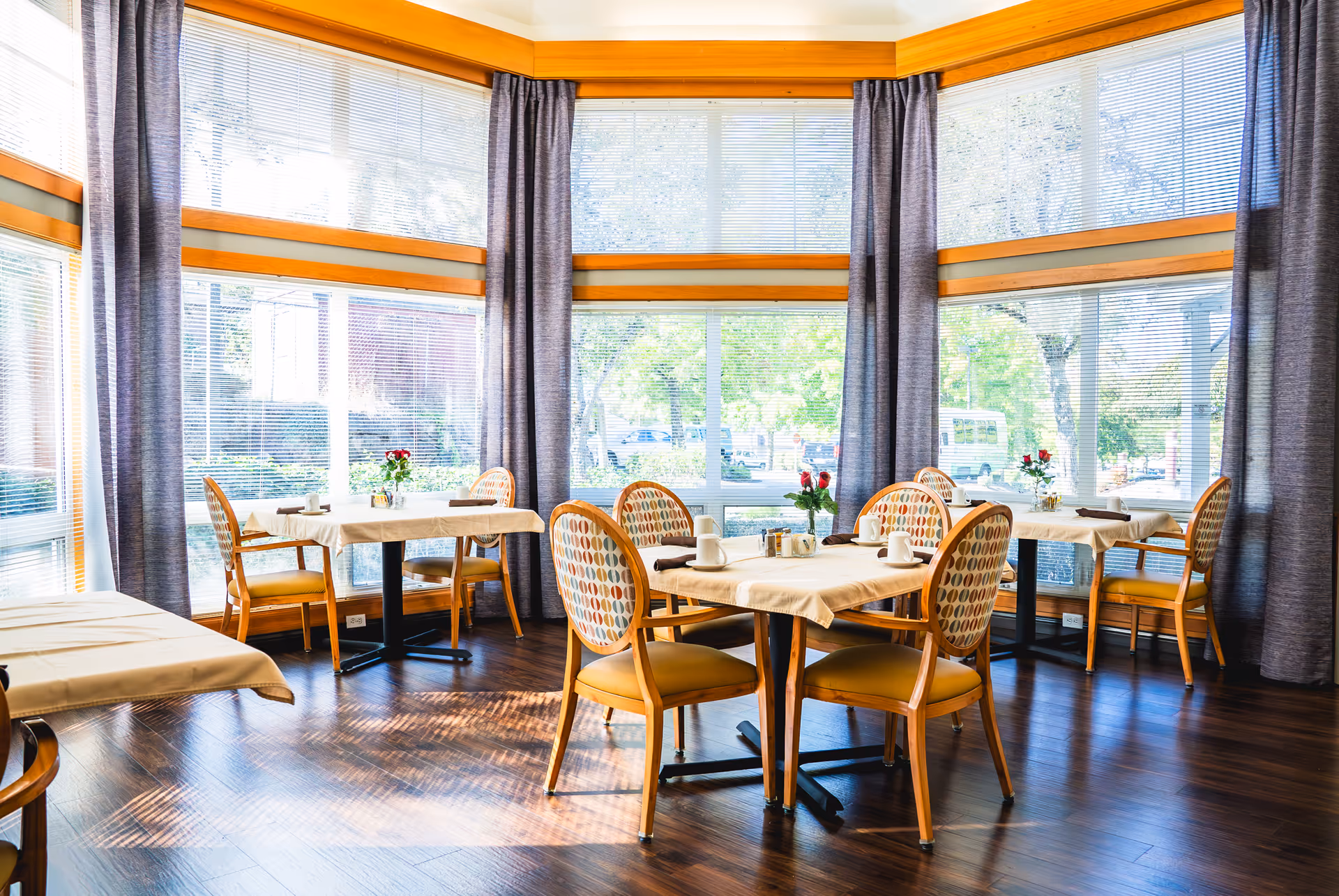 Bright dining room with large windows covered by blinds and gray curtains, featuring multiple tables with beige tablecloths, each set with chairs, coffee cups, and small flower vases with red roses.