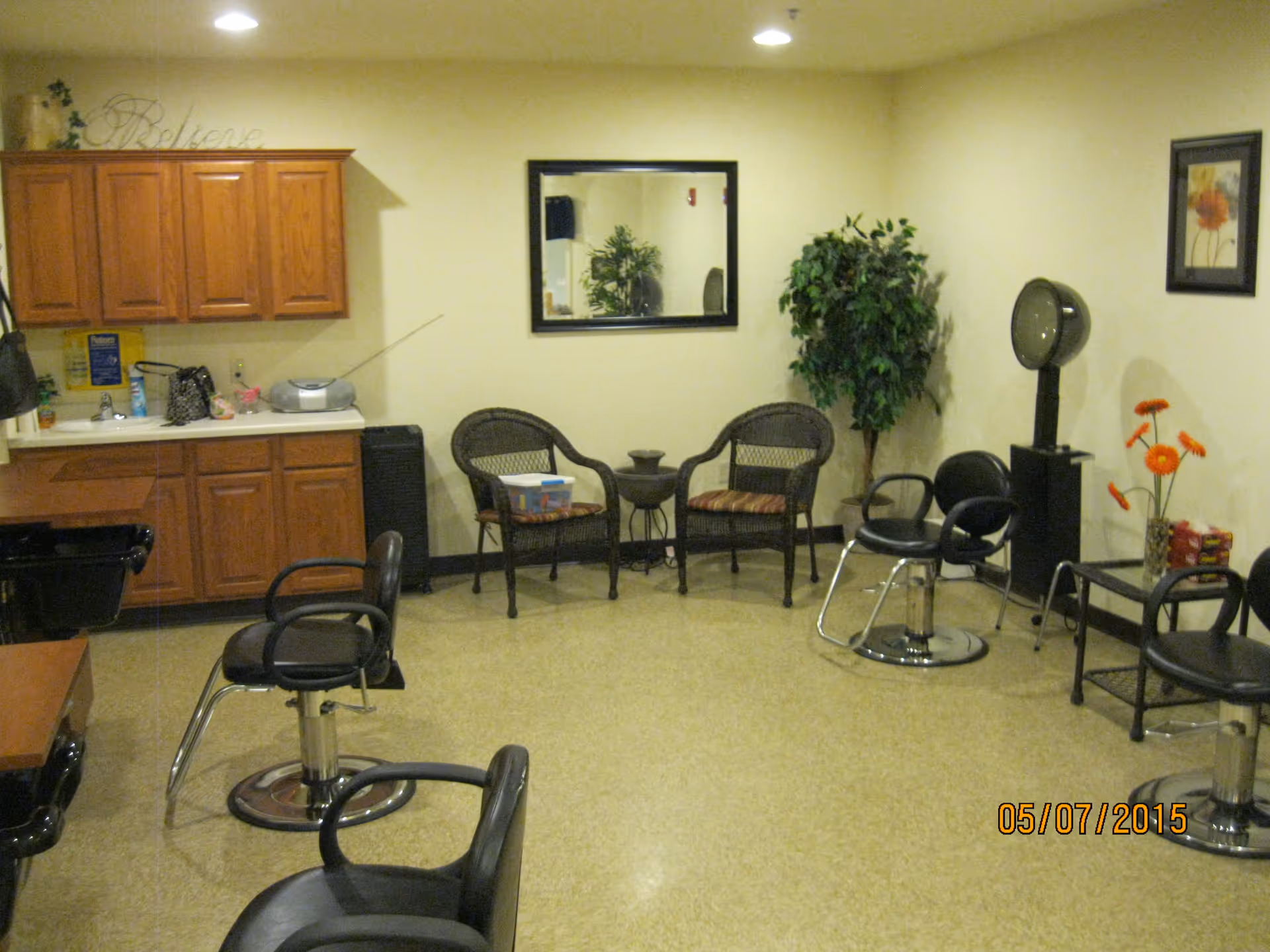 Interior room with salon chairs, a hair dryer, a small sink and cabinet area, two wicker chairs with a small table between them, a potted plant, and wall decorations including a mirror and framed artwork.