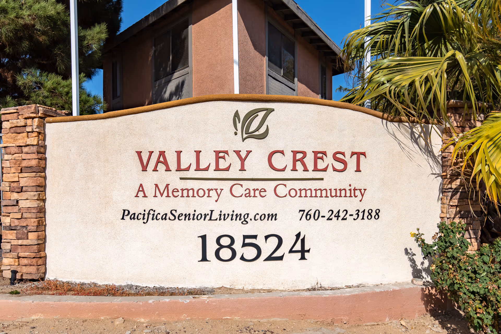 Stone and stucco sign for Valley Crest Memory Care Community with the website PacificaSeniorLiving.com and phone number 760-242-3188, set against a backdrop of a building, trees, and blue sky.