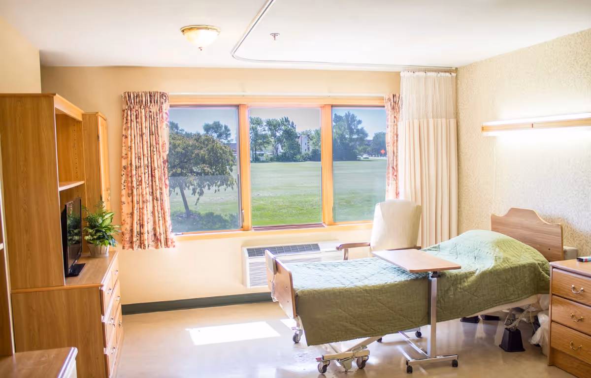 A bright and clean bedroom in a senior living facility with a hospital-style bed covered in a green quilt, a wooden headboard, a beige armchair, a wooden dresser with a small TV and a potted plant, floral curtains on the window, and a view of a green lawn and trees outside.