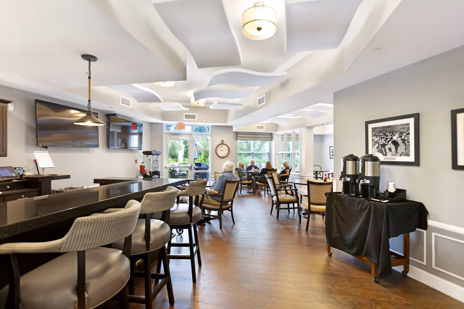 Interior view of a common area in an assisted living facility with a long counter and bar stools on the left, a coffee station on the right, and several elderly people seated at tables near large windows in the background. The room has modern ceiling lights and a wall-mounted TV.
