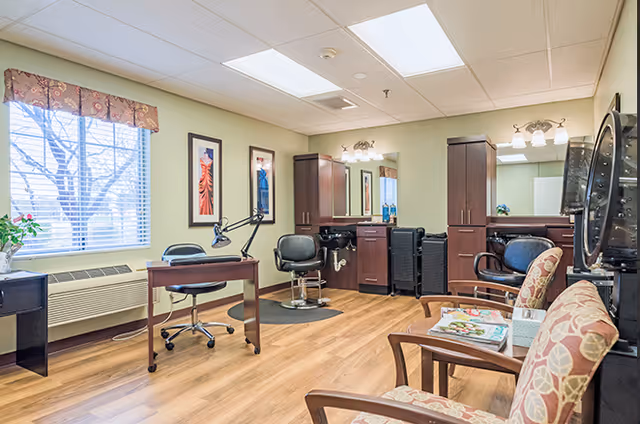 Interior of a senior living facility salon area with wooden flooring, two styling chairs in front of mirrors and cabinets, a desk with a lamp, and a seating area with patterned armchairs and magazines. A window with blinds and a floral valance lets in natural light.
