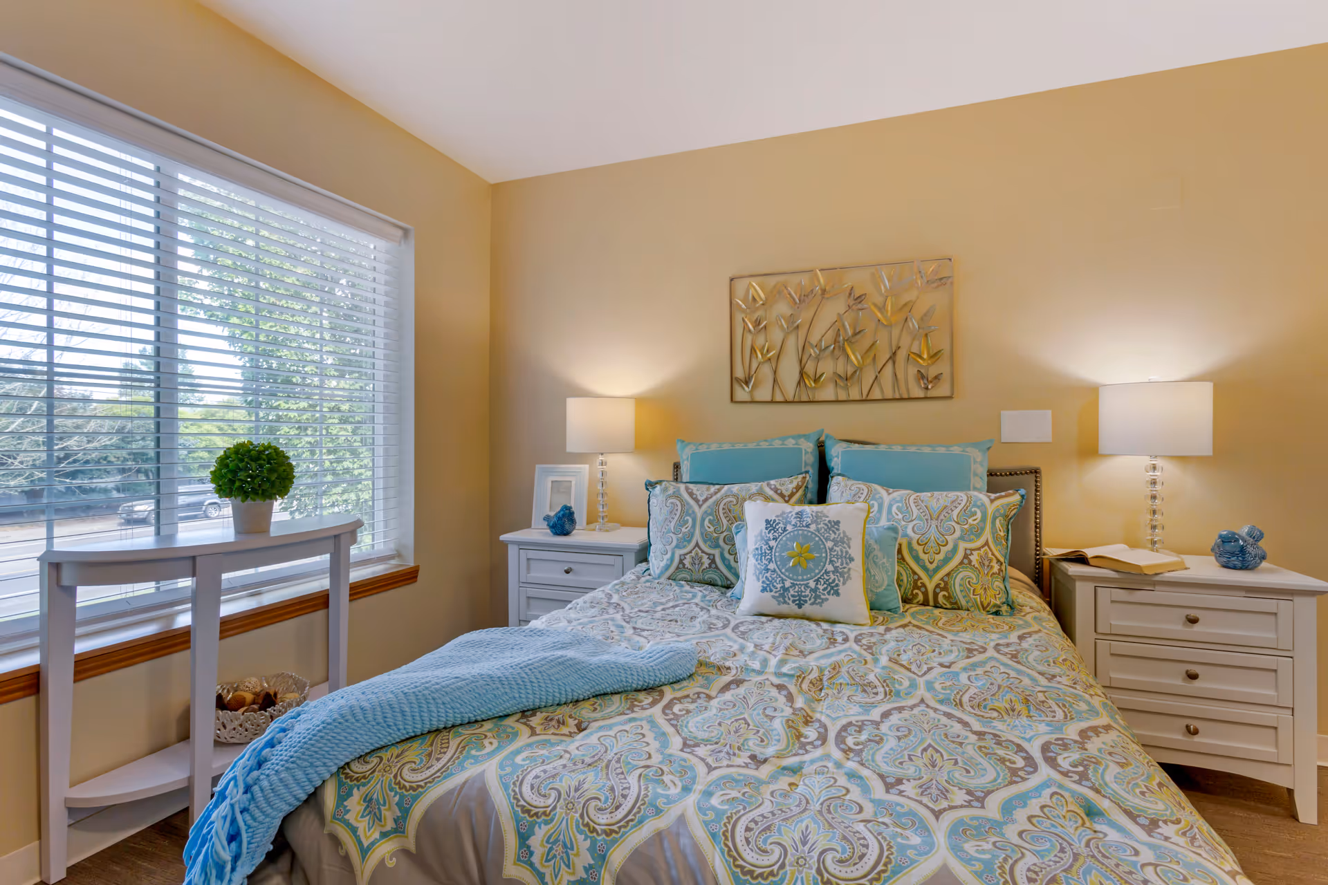 A cozy bedroom with a bed covered in a patterned blue, green, and beige comforter and multiple decorative pillows. There are two white nightstands on either side of the bed, each with a lamp and decorative items. A window with white blinds is on the left side, letting in natural light. The walls are painted a warm beige color, and a metal wall art piece featuring flowers hangs above the bed.