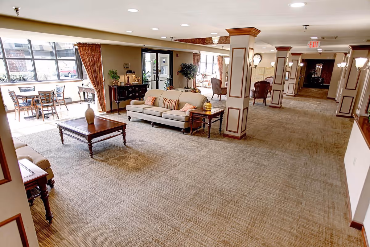 A spacious and well-lit common area in a retirement facility featuring a beige sofa with decorative pillows, wooden coffee and side tables, several chairs, and large windows with curtains allowing natural light to fill the room. The area has carpeted floors, decorative columns, wall sconces, and an exit sign in the background.
