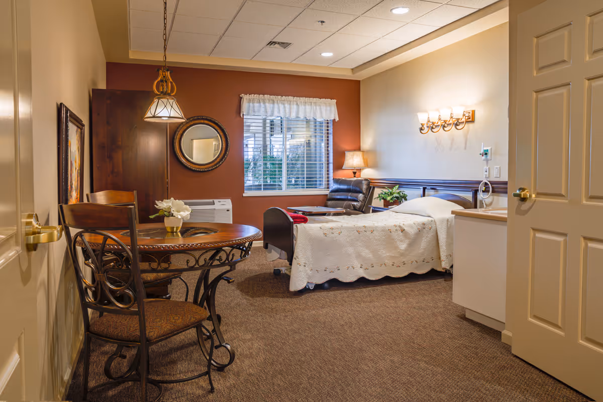 A cozy senior living room with a single bed covered in a white quilt, a brown leather recliner chair, a round wooden table with two chairs, a window with white blinds and a valance, a wall-mounted light fixture, a table lamp, and decorative wall art.