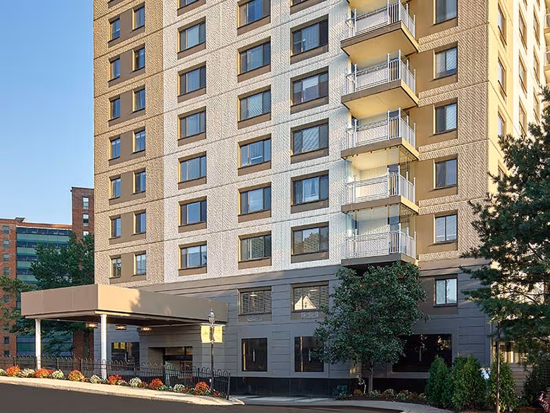 Exterior view of a multi-story senior living facility building with balconies, windows, and a covered entrance. There are trees and flower beds near the entrance, and other buildings are visible in the background under a clear blue sky.
