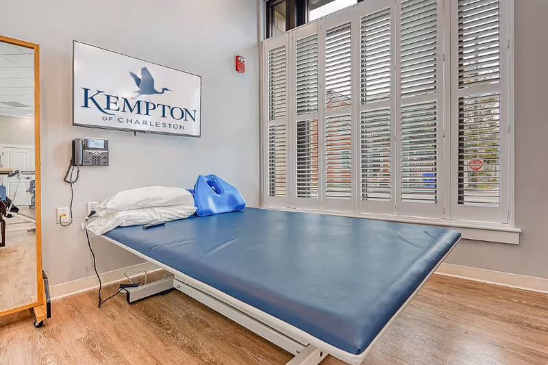 A therapy/exam room with a blue padded treatment table, pillows, a wall phone, and a 'KEMPTON of CHARLESTON' sign beside tall shuttered windows.