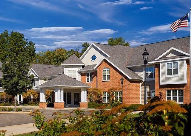 Exterior view of a senior living facility building with red brick and white siding, featuring a covered entrance, multiple windows, a lamppost, trees, and an American flag flying on a flagpole under a partly cloudy blue sky.