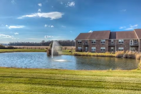 A two-story brick building next to a pond with a water fountain in the center, surrounded by green grass and open fields under a blue sky with scattered clouds.