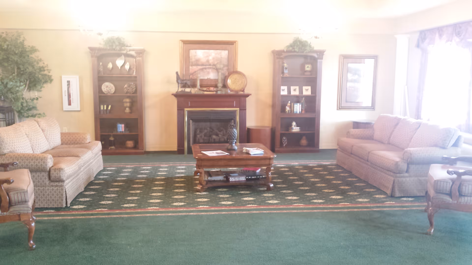 A cozy living room with two beige patterned sofas facing each other across a wooden coffee table on a green carpet with a floral pattern. The room features a fireplace with a decorative mantle, flanked by two wooden bookshelves filled with various decorative items. There are framed pictures on the walls and a large window with curtains letting in natural light.