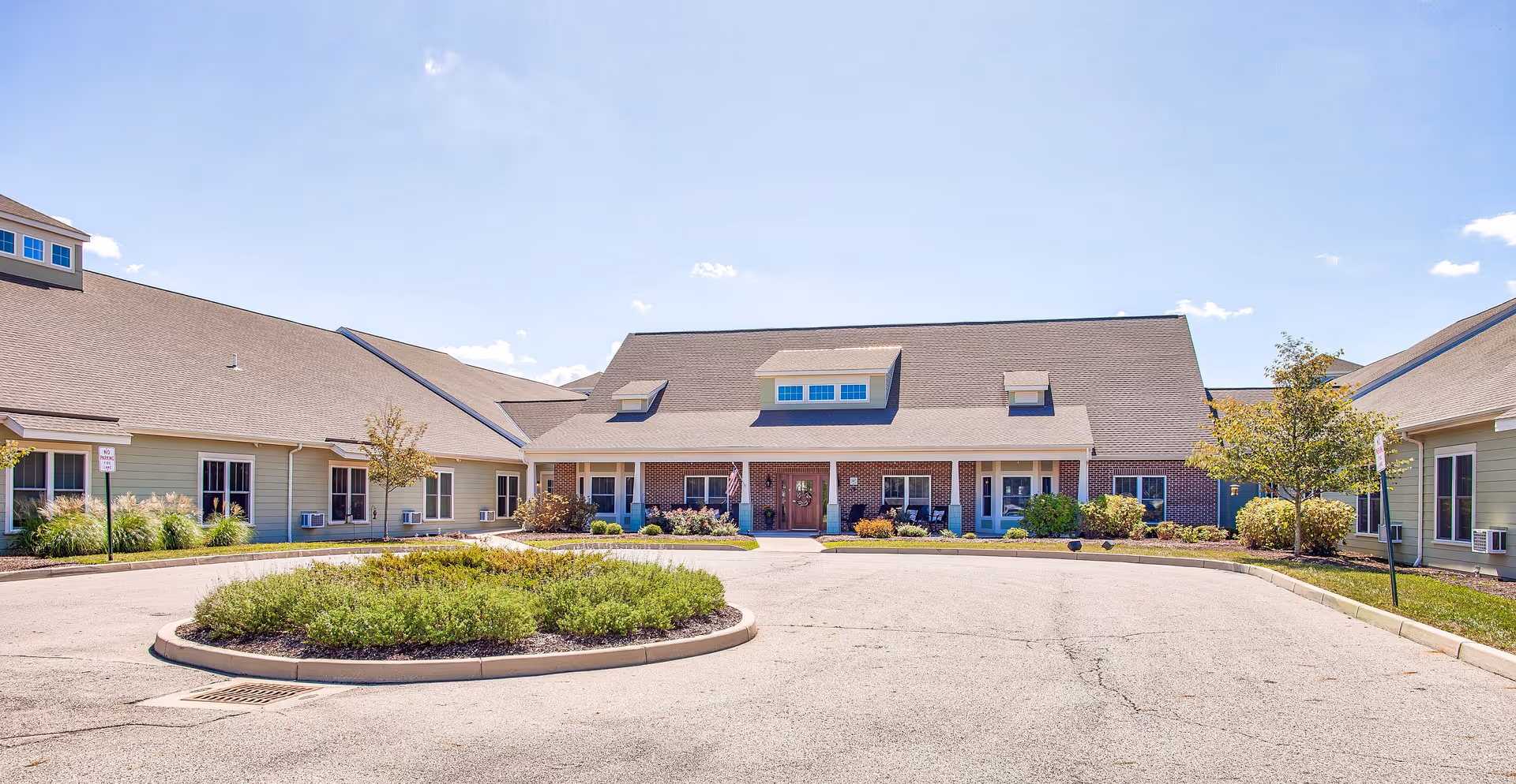 Front exterior view of a senior living facility with a circular driveway and landscaped greenery in the center. The building has a combination of brick and siding with multiple windows and a large roof with dormer windows under a clear blue sky.