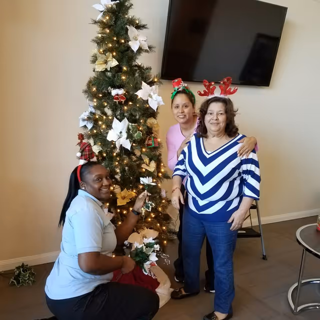 Three women smiling while decorating a Christmas tree in a communal living area.