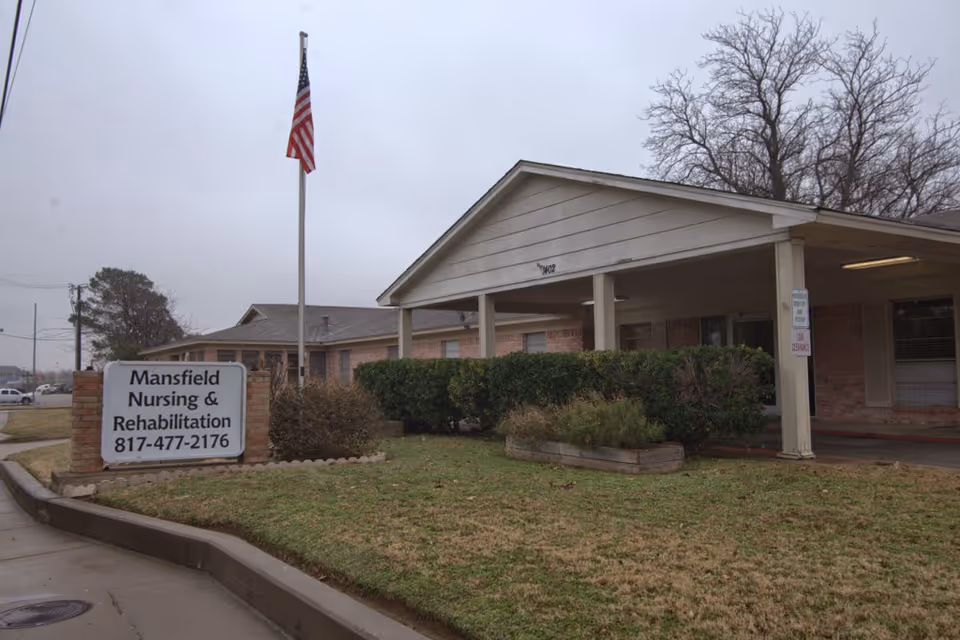 Exterior view of Mansfield Nursing & Rehabilitation Center showing a single-story brick building with a covered entrance, an American flag on a flagpole, and a sign displaying the facility's name and phone number. The surrounding area includes grass, bushes, and a sidewalk.