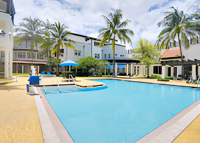 Outdoor swimming pool surrounded by a paved deck with blue lounge chairs and umbrellas. Palm trees and a multi-story white building with large windows are visible in the background under a partly cloudy sky.