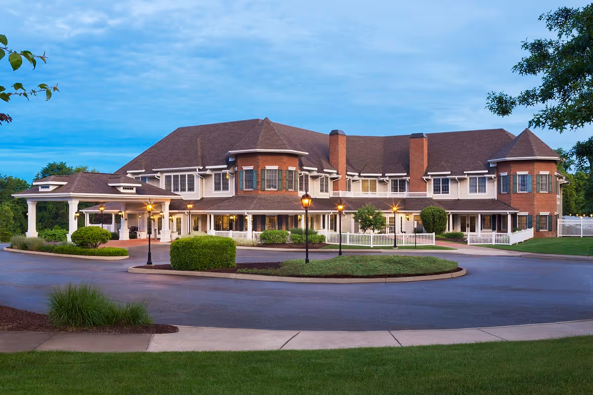 Exterior view of a large senior living facility building with a circular driveway, well-maintained landscaping, and multiple windows. The building has a combination of brick and siding with a covered entrance and several lamp posts lighting the driveway area.