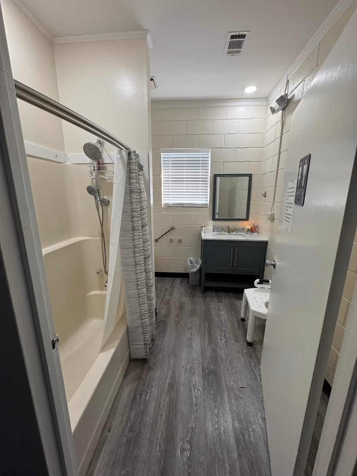 Interior view of a bathroom with a bathtub and shower on the left side, a shower curtain partially drawn, a window with blinds on the back wall, a vanity with a sink and mirror, a small trash can, and a white plastic shower chair on the right side near the door.