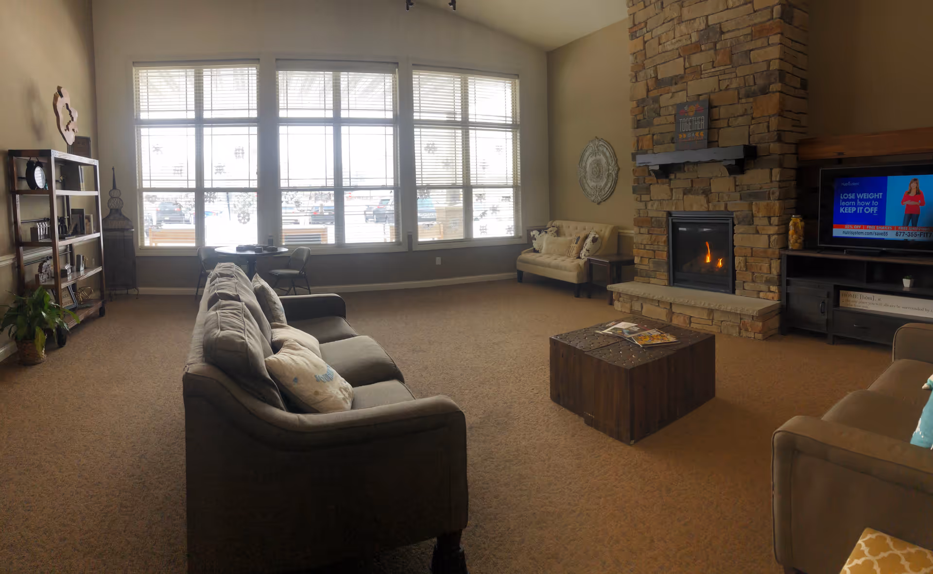A cozy living room with a large window letting in natural light, a stone fireplace with a fire burning, a TV on a stand, two sofas with pillows, a small loveseat, a wooden coffee table with magazines, a bookshelf with decorative items, and a small table with two chairs near the window.