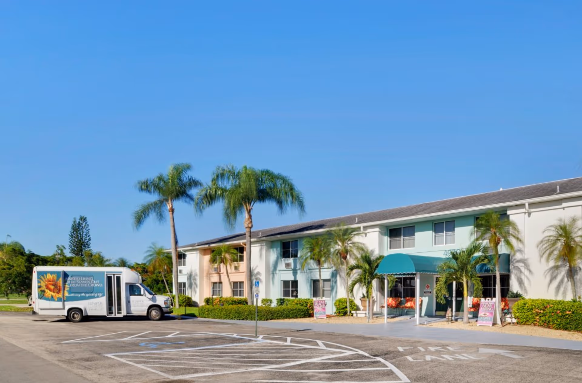 Exterior view of a two-story senior living facility building with pastel-colored walls and a teal awning over the entrance. Palm trees and shrubs are planted around the building. A white shuttle bus with a sunflower image and text is parked in the lot near the building. The parking lot has marked spaces including a handicapped spot and a fire lane.