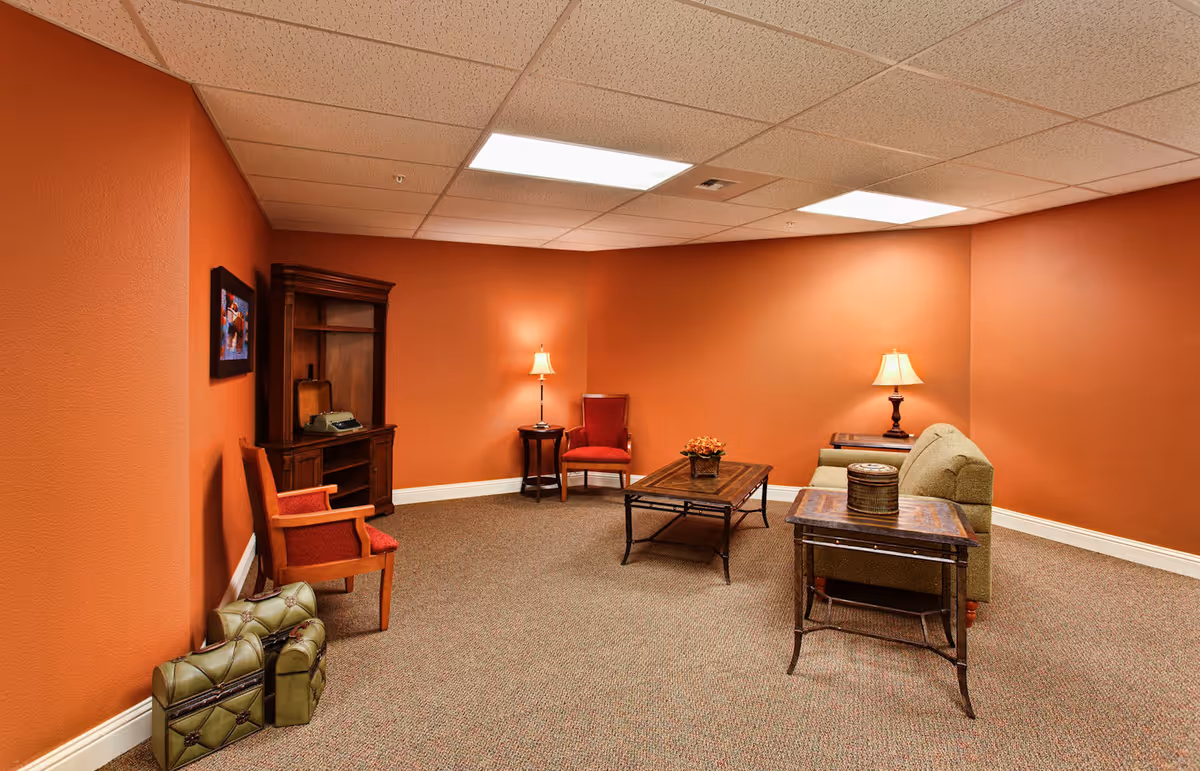 A cozy living room with orange walls and carpeted floor, featuring a green sofa, two red armchairs, two wooden side tables with lamps, a wooden coffee table with a small plant, a wooden cabinet with a vintage typewriter, and a framed picture on the wall.