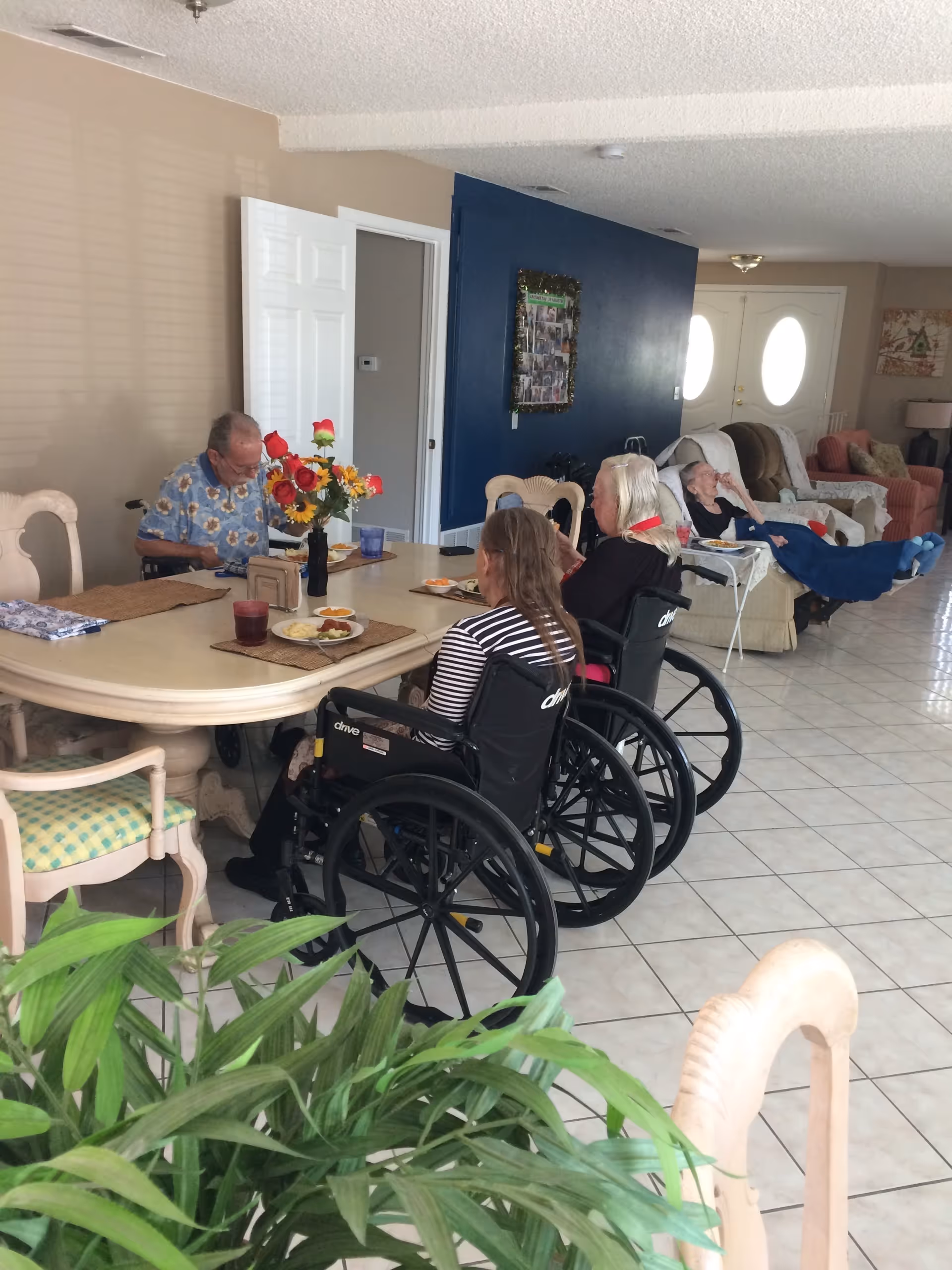 A group of elderly individuals, some in wheelchairs, sitting around a dining table with plates of food in a bright room. There are flowers in a vase on the table, and a few people are seated on couches in the background. The room has tiled floors, a blue accent wall, and double doors with oval windows.